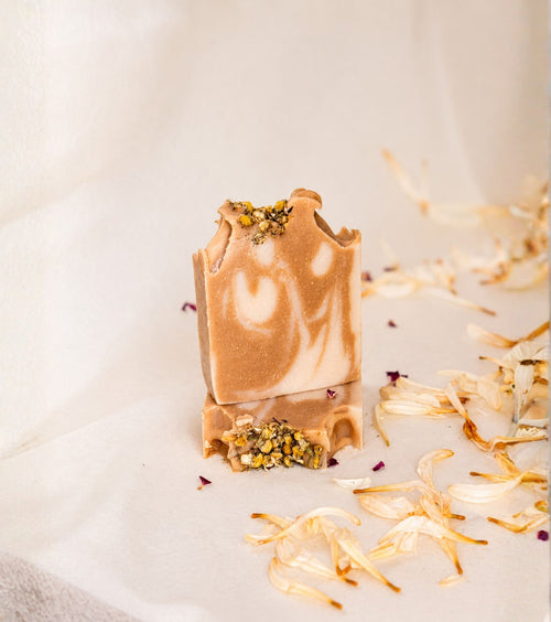 Natural handmade botanical soap with chamomile flowers, creamy ivory and caramel swirls, photographed on soft neutral fabric with scattered flower petals for a luxury skincare presentation.