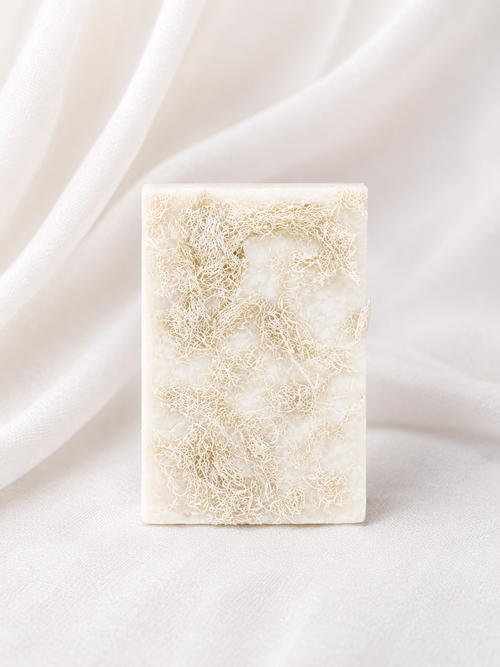 Natural loofah soap bar with embedded loofah fibers, handmade for gentle exfoliation and daily cleansing, displayed on a soft neutral fabric background.