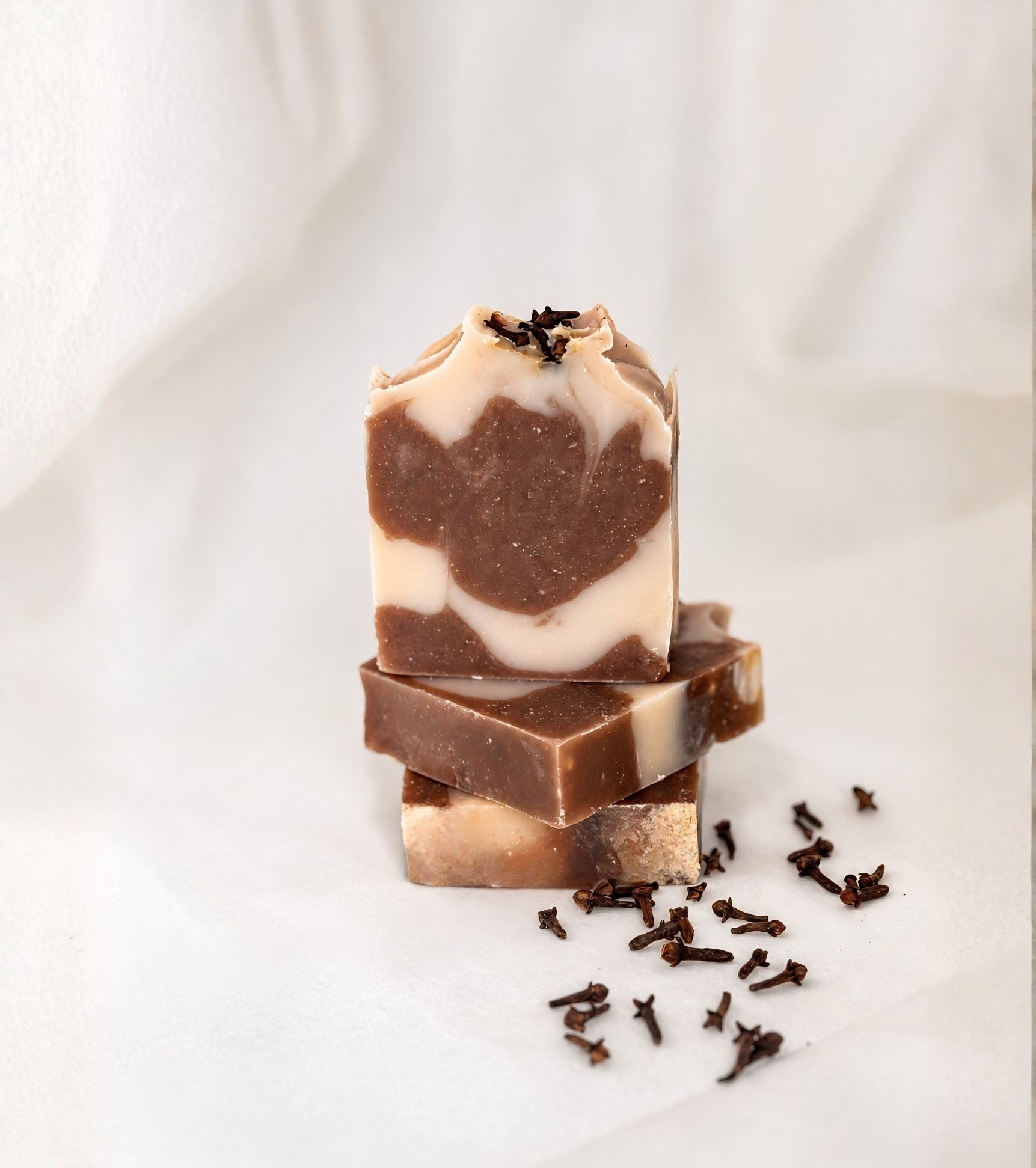 Three stacked bars of handmade clove bud soap with creamy beige and rich brown swirls, styled on soft draped fabric with scattered dried cloves for a warm, natural skincare aesthetic.